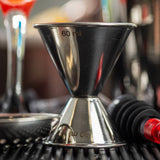 A close-up of a stainless steel jigger measuring cup, showing 60 ml and 30 ml markings. The 1 oz 2 oz jigger sits on a black bar mat with cocktail tools and blurred drinks in the shiny, reflective background.