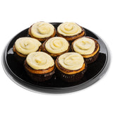 Seven cinnamon rolls topped with swirls of cream cheese frosting are arranged in a circular pattern on a shiny Sierra Collection Serving Platter by TableCraft Products, viewed from above against a white background. The golden brown rolls sit in dark paper wrappers.