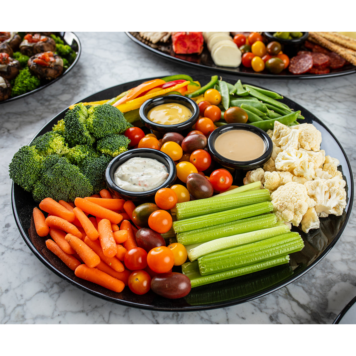 A black round platter from the Sierra Collection is filled with fresh vegetables—broccoli, carrots, cherry tomatoes, celery, cauliflower, bell peppers, and snap peas—with three dips in the center. The melamine serving platter rests on a marble surface.