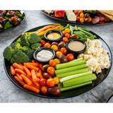 A black round platter from the Sierra Collection is filled with fresh vegetables—broccoli, carrots, cherry tomatoes, celery, cauliflower, bell peppers, and snap peas—with three dips in the center. The melamine serving platter rests on a marble surface.