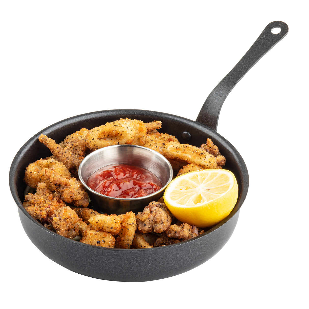 A black mini serving fry pan contains crispy fried chicken pieces, a metal cup of red dipping sauce (likely cocktail or marinara), and a halved lemon, cut side up, all on a white background.