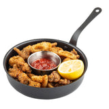 A black mini serving fry pan contains crispy fried chicken pieces, a metal cup of red dipping sauce (likely cocktail or marinara), and a halved lemon, cut side up, all on a white background.