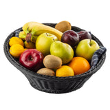 A black, round polypropylene basket, like the TableCraft 12129, is filled with assorted fruits—bananas, apples, pears, kiwis, plums, oranges, lemons, and a lime. A dark blue and white striped cloth peeks through. The background is plain white.