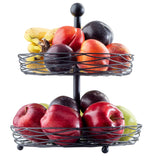 A two-tiered black steel display basket holds bananas, plums, nectarines, red apples, green apples, and two dark purple fruits. The fresh, colorful fruits are beautifully arranged against a white background.