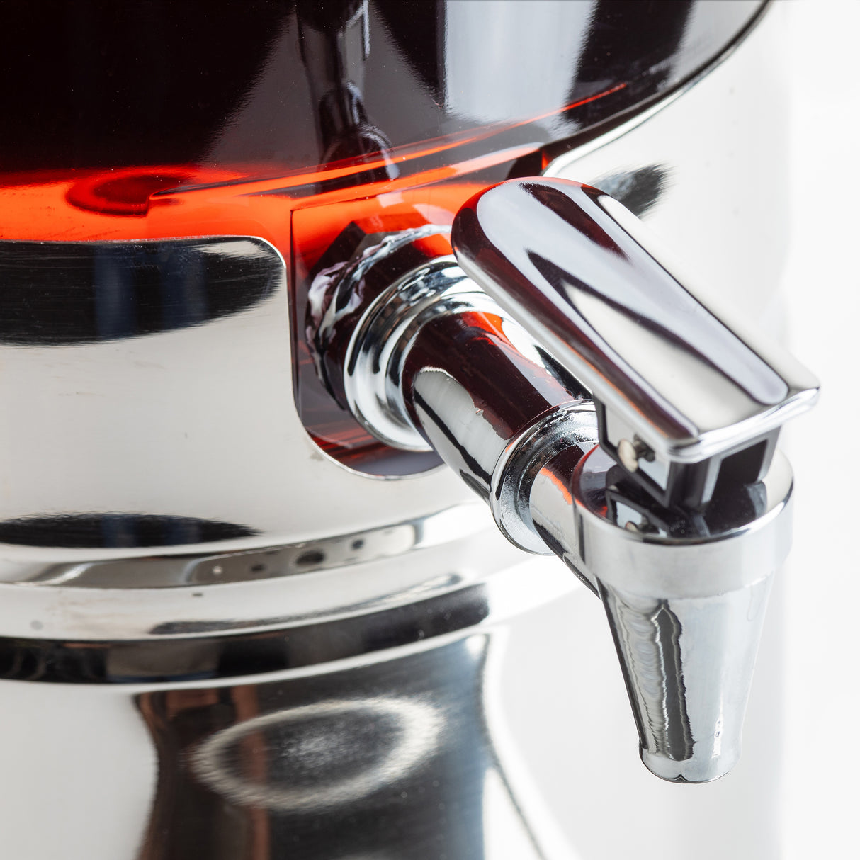 Close-up of a shiny chrome spigot with lever handle on a 2 gallon polycarbonate beverage dispenser. Red liquid reflects on the polished metal, while the softly blurred white background highlights its clean, modern appeal.