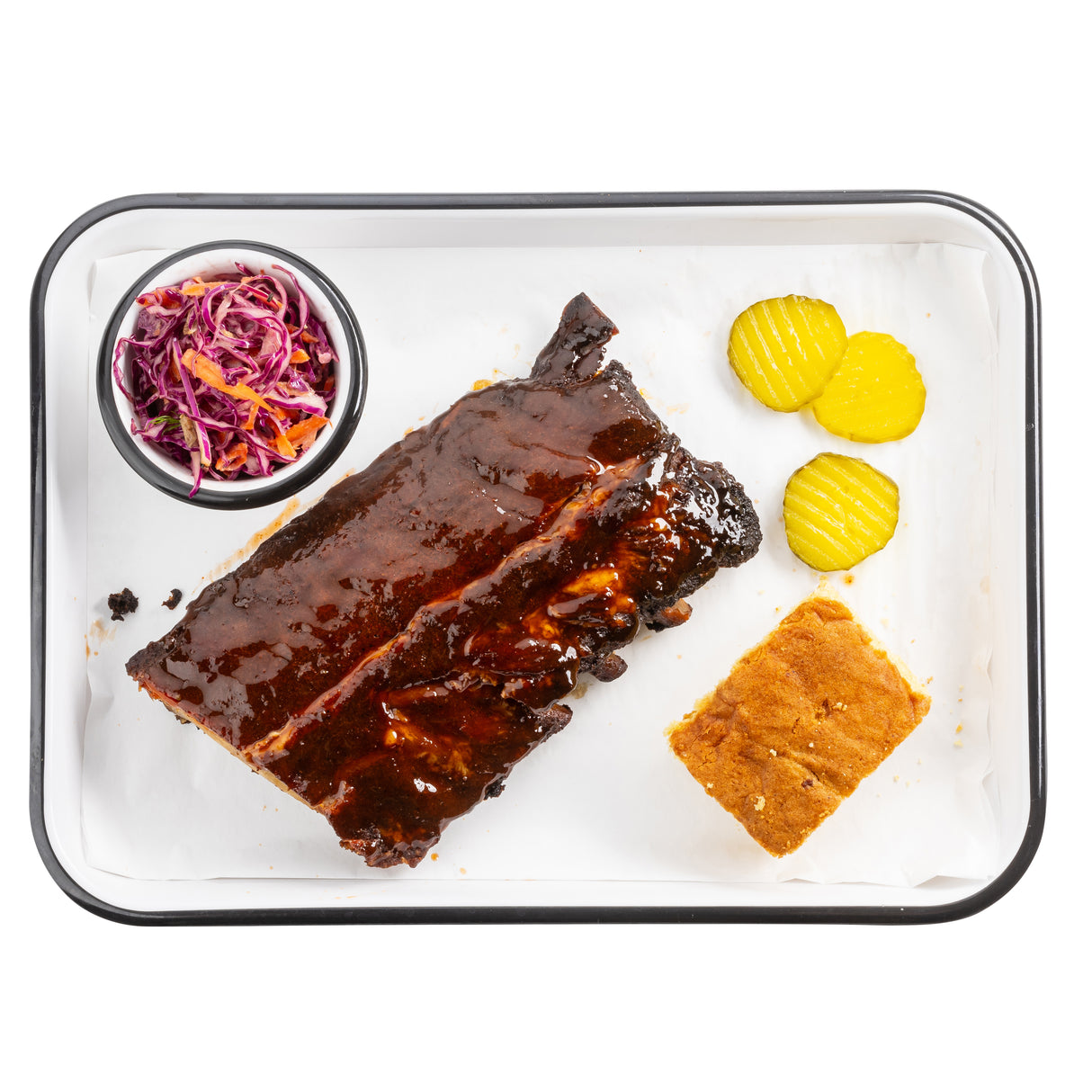 A Melamine serving tray holds saucy barbecue ribs, cornbread, pickle slices, and colorful coleslaw—all neatly arranged on white parchment paper with a white background.