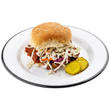 A sesame seed bun sandwich filled with shredded barbecue meat and coleslaw sits on a TableCraft Products Canteen Collection Plate with a thin black rim. Two crinkle-cut pickle slices are beside the sandwich. The background is white.