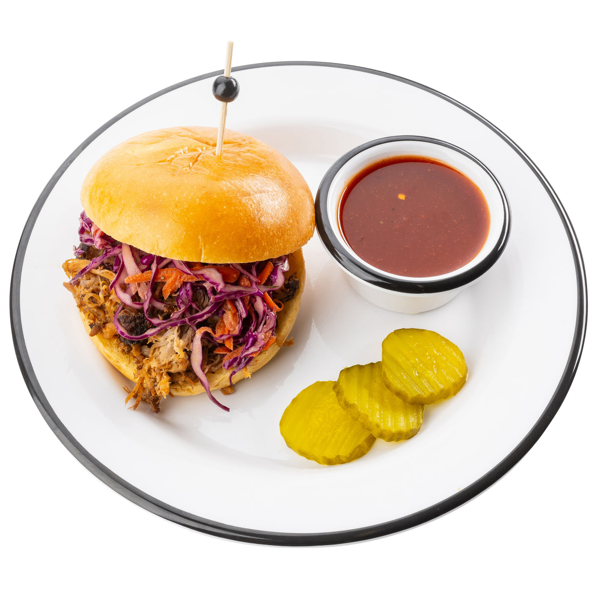 A white TableCraft Canteen Collection Plate holds a pulled pork sandwich with coleslaw on a brioche bun, secured by a toothpick. Three pickle slices and a small metal cup of reddish-brown barbecue sauce sit beside it, all against a white background.