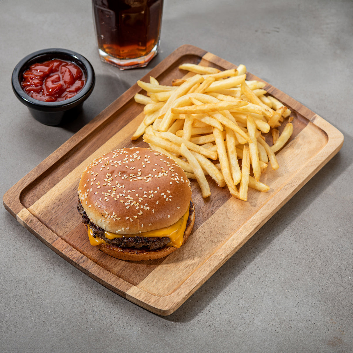 A cheeseburger with a sesame seed bun, melted cheese, and beef patty sits on a rectangular serving board next to golden French fries. Beside the board are a cup of ketchup and a glass of dark soda on a gray tabletop.