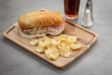 A wood serving plate holds a sub sandwich with deli meats, cheese, shredded lettuce, and a side of potato chips. Behind the tray is a glass of iced tea and salt and pepper shakers on a plain, light-colored tabletop.