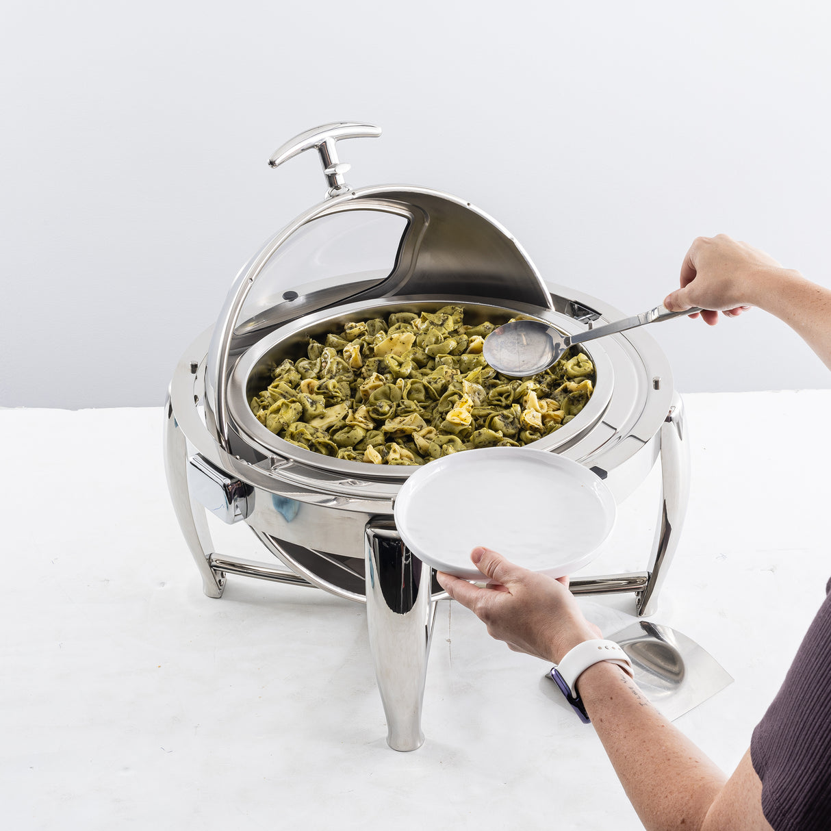 A person holds a white plate and uses a serving spoon to scoop pesto tortellini from a 6 qt. stainless steel chafer with a dripless roll top lid, sitting on a white table with a modern, polished design.