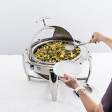 A person holds a white plate and uses a serving spoon to scoop pesto tortellini from a 6 qt. stainless steel chafer with a dripless roll top lid, sitting on a white table with a modern, polished design.