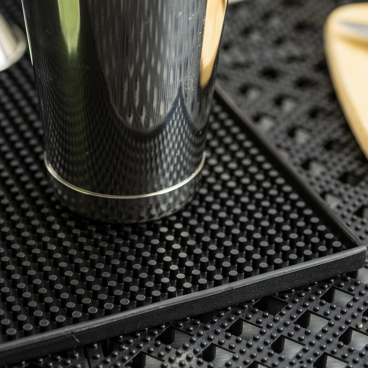 A close-up of a metallic cocktail shaker resting on a black rubber service mat with small raised dots, placed on a perforated surface. Part of a wooden object is visible in the upper right corner. The mat is dishwasher safe for easy cleaning.