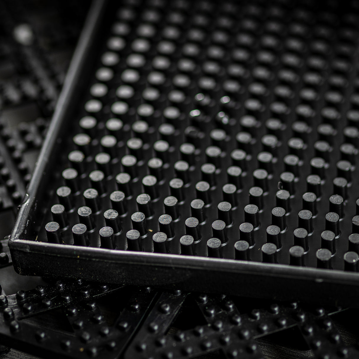 Close-up of a black rubber tray filled with evenly spaced vertical pegs or cylinders. The tray, likely a service mat, has a rectangular border and textured surfaces in the background. Lighting creates subtle reflections. Dishwasher safe for easy cleaning.