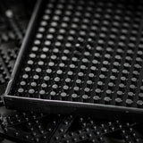 Close-up of a black rubber tray filled with evenly spaced vertical pegs or cylinders. The tray, likely a service mat, has a rectangular border and textured surfaces in the background. Lighting creates subtle reflections. Dishwasher safe for easy cleaning.