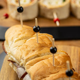 A close-up of a sandwich cut into sections with TableCraft Products Ball Picks, each topped with black olives. The sandwich sits on a wooden board, with blurred pinwheel appetizers on bamboo and pinewood skewers in the background. The bread looks soft and golden-brown.