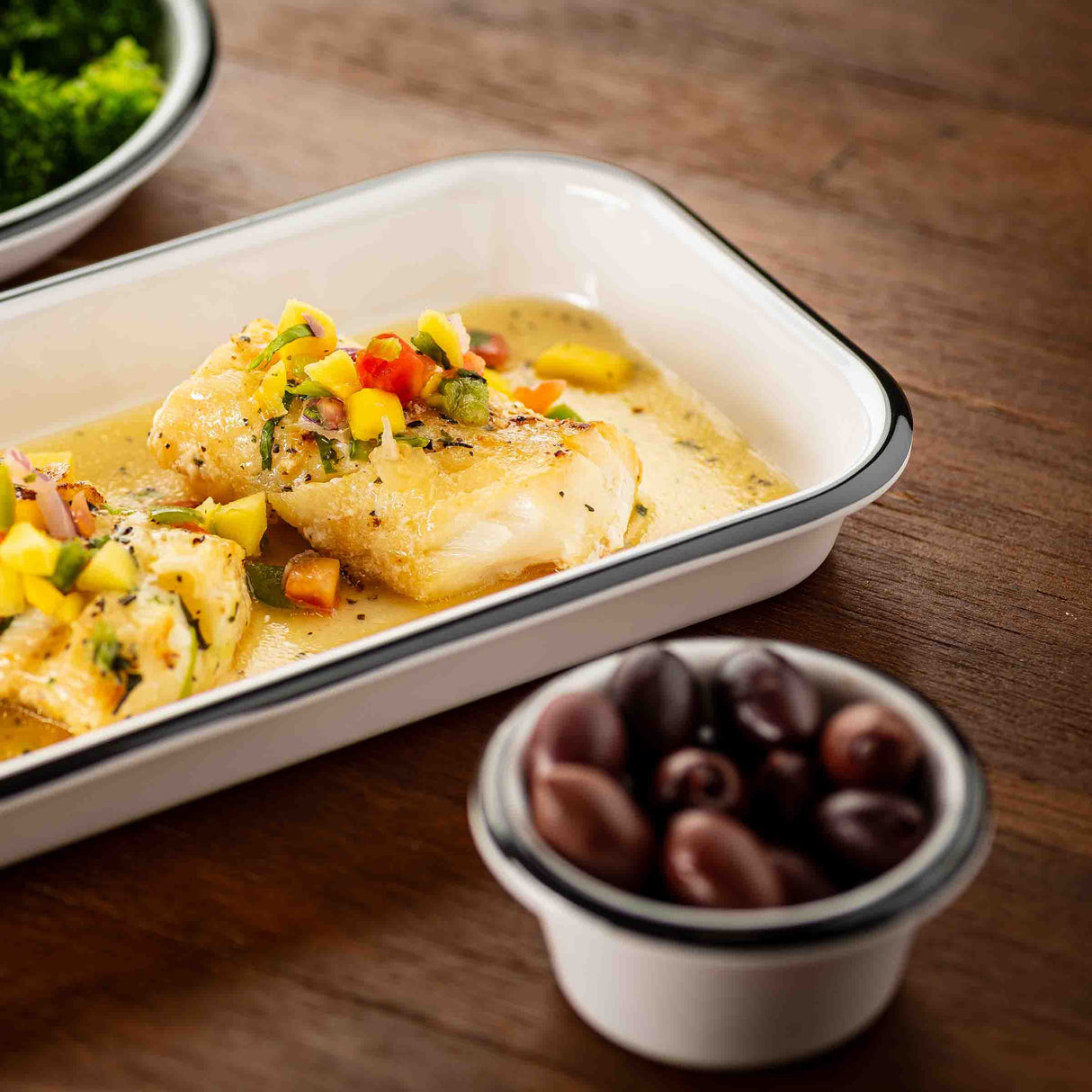 A white enamel dish, possibly TableCraft, contains two pieces of fish topped with diced yellow and red vegetables in a creamy sauce. In the foreground, a small ramekin holds black olives. Green vegetables can be seen in the background on a wooden table.