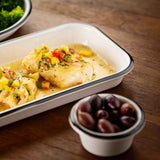 A white enamel dish, possibly TableCraft, contains two pieces of fish topped with diced yellow and red vegetables in a creamy sauce. In the foreground, a small ramekin holds black olives. Green vegetables can be seen in the background on a wooden table.