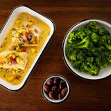 A meal on a wooden table: two pieces of grilled fish with mango salsa and yellow sauce served in a rectangular melamine dish, a bowl of steamed broccoli, and black olives presented in a small ramekin.