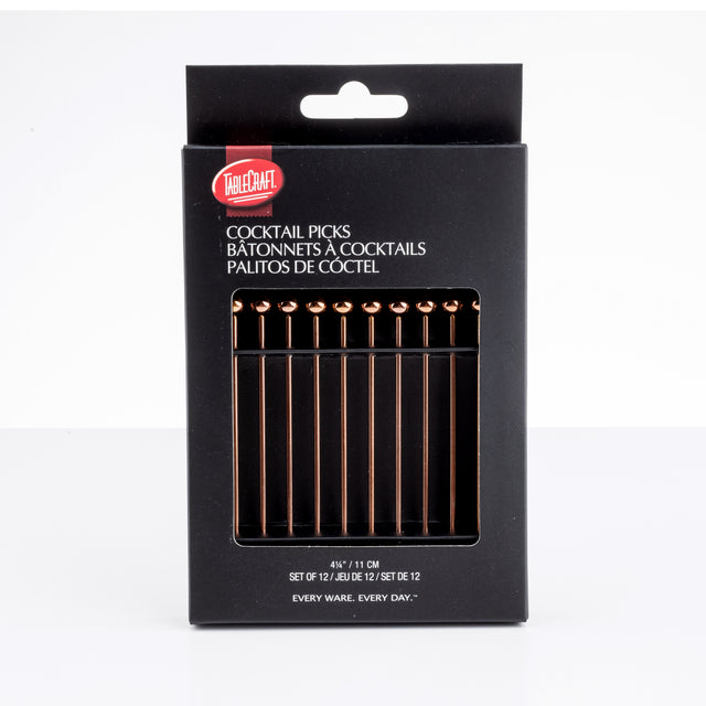 A black box labeled “TableCraft Cocktail Picks” displays twelve gold-toned stainless steel picks inside. The packaging features white text in English, French, and Spanish, plus a window revealing the picks arranged neatly in a row.