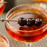 A close-up of a cocktail glass filled with a reddish-brown drink, garnished with three glossy dark cherries skewered on a gunmetal stainless steel cocktail pick. The delicate rim and slight frost suggest the drink is refreshingly cold.