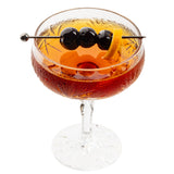 A coupe glass filled with a reddish-brown cocktail is garnished with three dark cherries and a twist of orange peel, both skewered on a sleek stainless steel cocktail pick. The etched glass, covered in condensation, stands out against a white background.