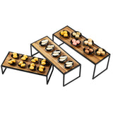 Three rectangular Acacia wood risers with black powder coated steel frames are arranged at different heights. Each stand holds several black plates, displaying assorted pastries including macarons and cream-filled buns, neatly organized in rows. The background is white.