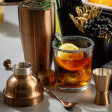 A glass of iced cocktail garnished with lemon slices and a sugared rim sits on a table. Surrounding it are bar kit essentials, including a stainless steel cocktail shaker, measuring jigger, spoon, and strainer. Stir sticks and picks rest in a black container nearby.