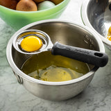 A metal bowl with separated egg whites sits on a marble counter. A TableCraft egg separator with a black handle holds a yolk above the bowl. Behind it, a green bowl contains brown and white eggs, with another metal bowl partially visible to the right.