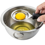 A hand holds a TableCraft black-handled egg separator over a stainless steel bowl. The separator cradles a yellow yolk as the white drips into the bowl, which features measurement markings and a convenient side handle.