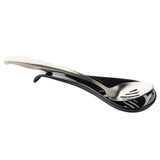 A stainless steel slotted spoon rests on a glossy black TableCraft spoon rest. The curved, elongated melamine holder is designed to catch drips from the spoon, whose handle extends over the edge while its bowl features three slots for draining.