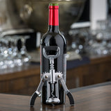 A bottle of red wine with a red and green foil cap stands on a wooden surface. A TableCraft Premium Winged Corkscrew is inserted into the cork, its arms angled downward. Blurred glassware and a metallic object are visible in the background.