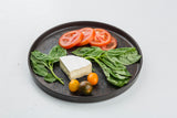 A dark Lunara Collection platter holds spinach leaves, three slices of tomato, a wedge of brie cheese, one red grape tomato, and two yellow grape tomatoes, all arranged neatly on a white background.
