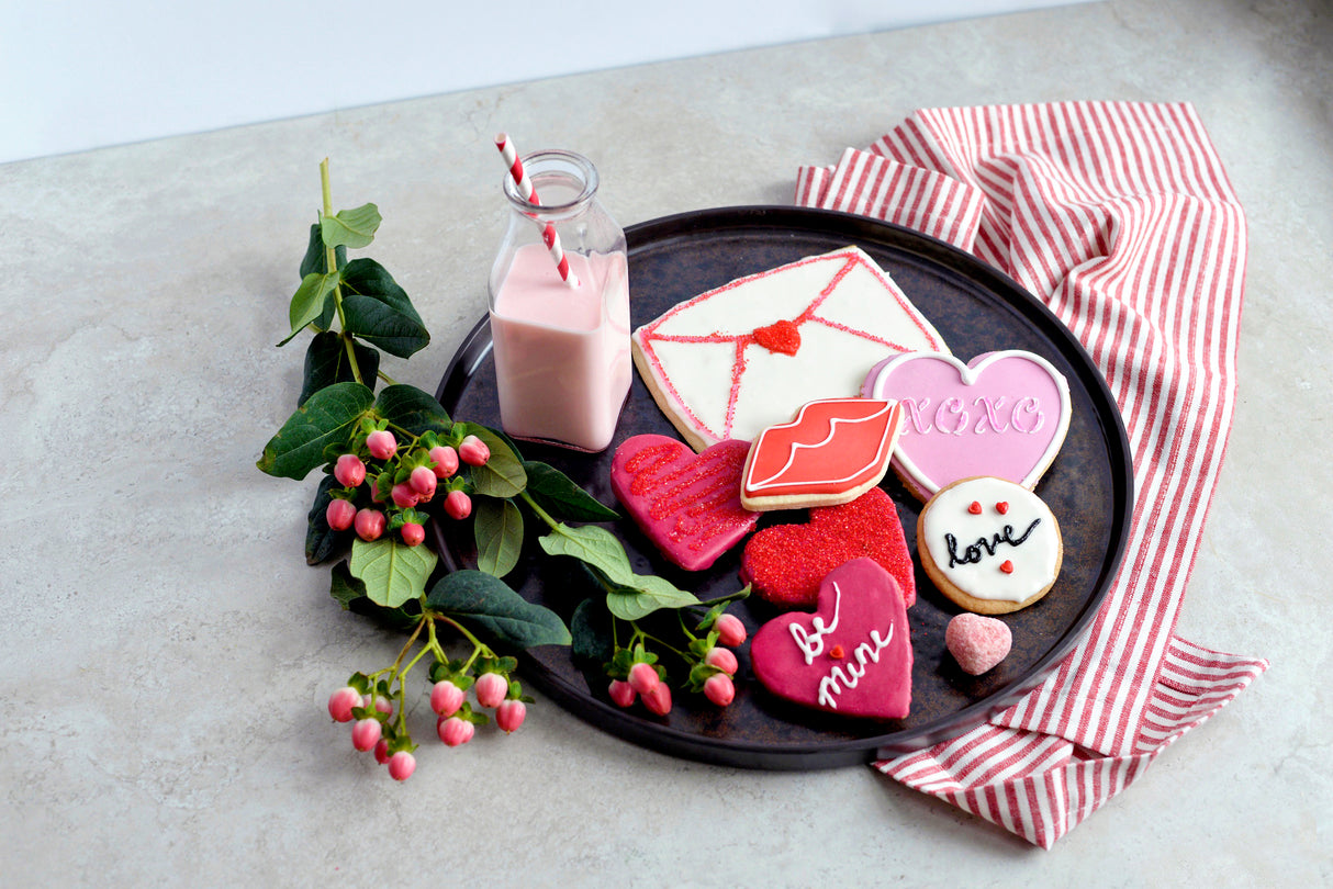 A dark TableCraft melamine platter holds heart-shaped Valentine cookies, a letter-shaped cookie, a pink “XOXO” cookie, a “love” cookie, a lips cookie, pink milk with straws, greenery with pink berries, and a red-striped cloth on a light surface.