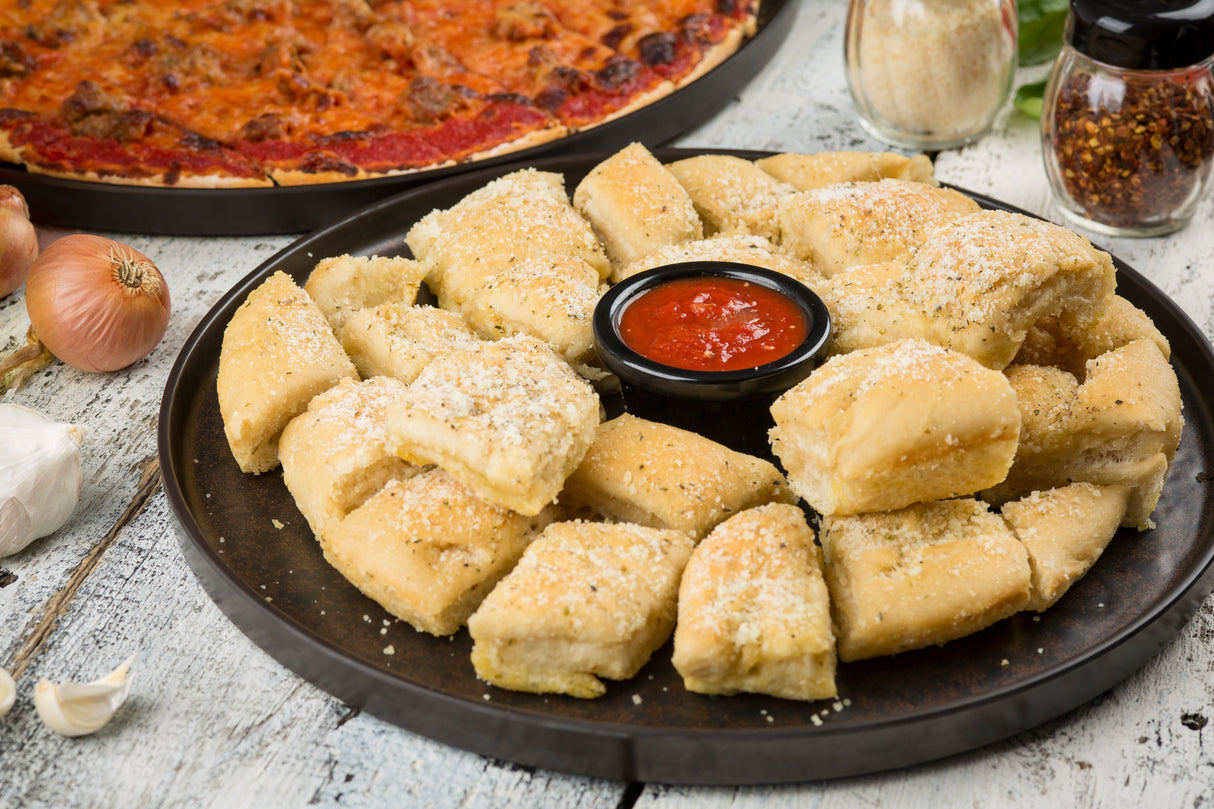 A round melamine platter from the TableCraft Lunara Collection holds golden, fluffy garlic bread bites sprinkled with parmesan cheese, arranged around a small cup of marinara sauce on a rustic white table.
