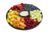 A round black melamine platter from the Lunara Collection by TableCraft holds sections of strawberries, blueberries, honeydew melon, raspberries, cantaloupe, and pineapple around a central bowl of creamy white dip. The fruit is neatly arranged on a white background.