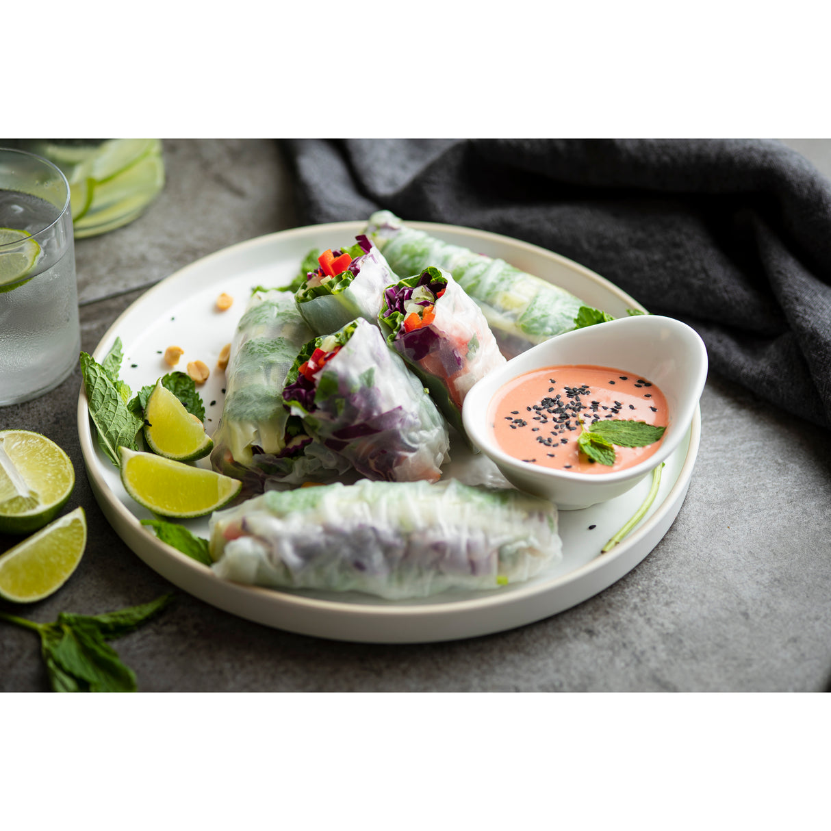 Four fresh vegetable spring rolls sit on a round white tray, with lime wedges, mint leaves, and chopped peanuts on the side. A small bowl of pink dipping sauce sprinkled with black sesame seeds completes this vibrant Europa Collection Serving Tray display.