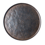 A round serving tray with a textured, uneven surface and mottled pattern. This ceramic platter features black and brown tones for a rustic, earthy look. The plain white background highlights the design of the plate.