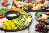 A colorful platter of fresh vegetables surrounds a bowl of creamy dip. Nearby trays, including a stylish melamine tray, offer cheese, crackers, breadsticks, grapes, cherry tomatoes, olives, and sliced meats on a rustic white surface.