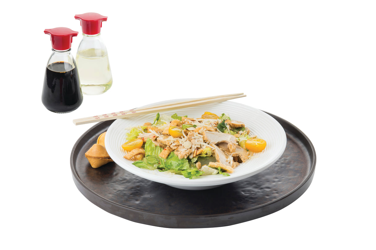 A white bowl of salad with chicken, lettuce, and yellow cherry tomatoes sits on a round serving tray with chopsticks. A fortune cookie is beside the bowl. Two glass bottles of soy sauce and vinegar are in the background against a white backdrop.