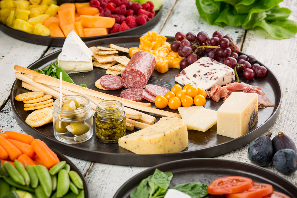 A round serving platter with assorted cheeses, cured meats, crackers, breadsticks, fresh grapes, berries, cherry tomatoes, figs, olives in a jar, and sliced vegetables—all arranged on a rustic white wooden table.
