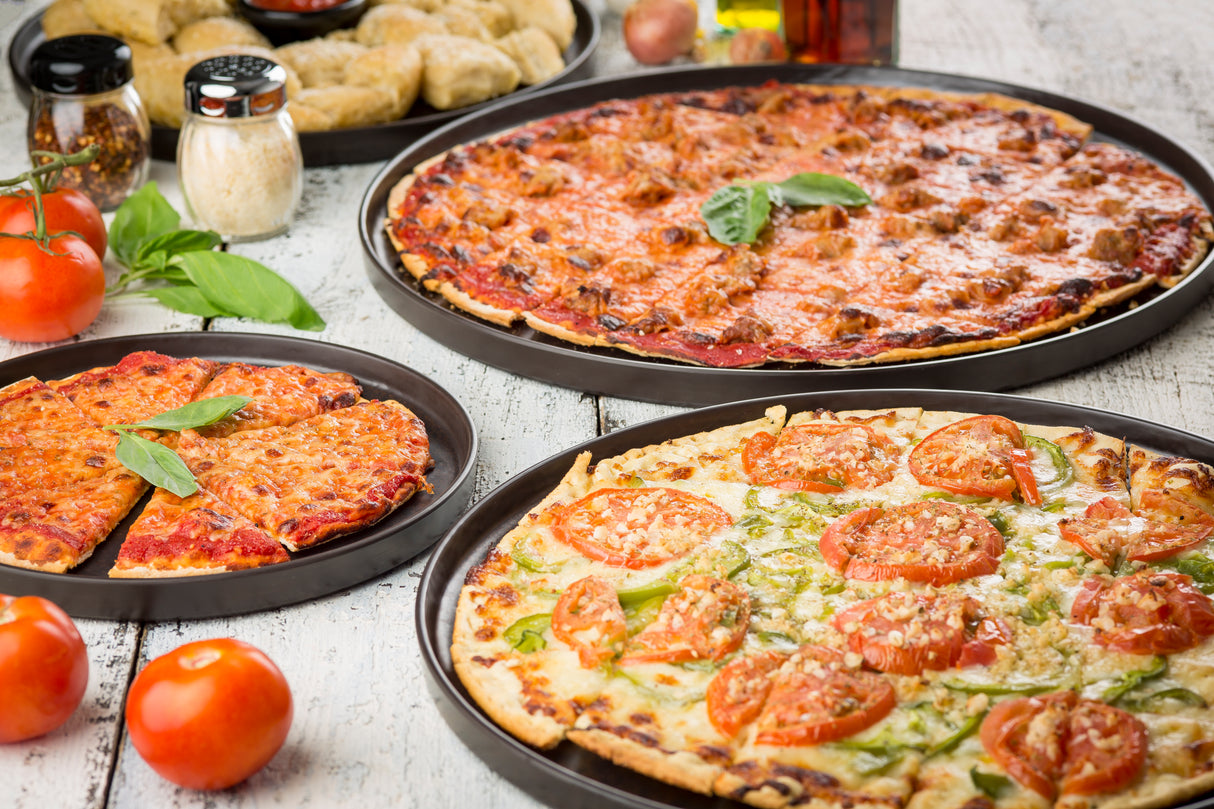 Three pizzas on metal trays sit on a rustic white table: sausage, plain cheese, and one with fresh tomato and basil. In the background is a Lunara Collection platter with breaded snacks, tomatoes, Parmesan, and condiments. Fresh basil adds color.