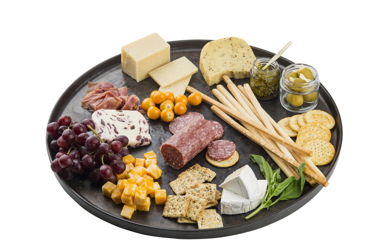 A round serving platter holds assorted cheeses, salami, prosciutto, crackers, breadsticks, grape tomatoes, green olives in a jar, a basil sprig, red grapes, and cubed cheese—all arranged neatly with small jars and a cheese knife. White background.