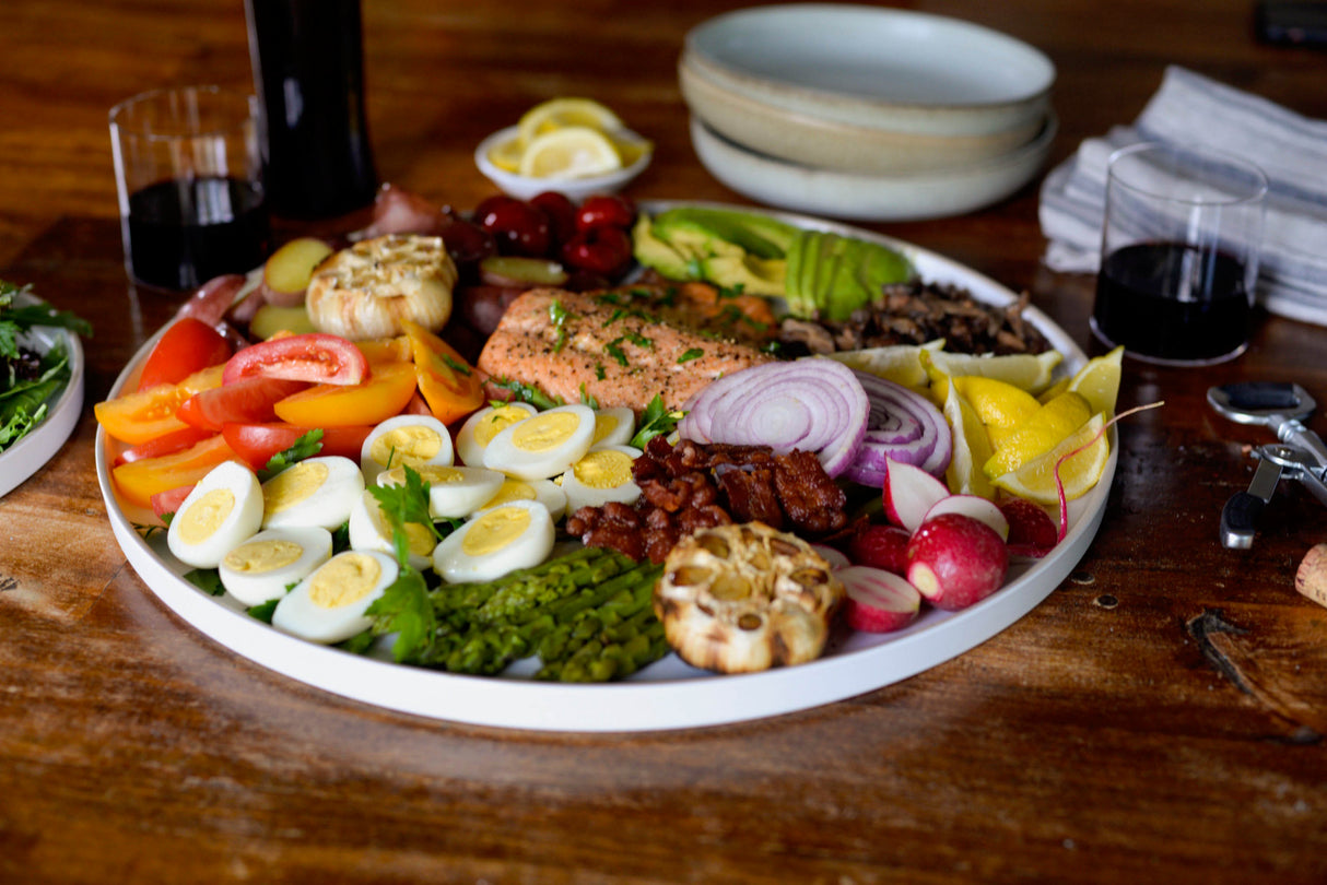 A large round melamine serving tray holds hard-boiled eggs, sliced tomatoes, asparagus, roasted garlic, radishes, lemon wedges, red onion, bacon, salmon, olives, pickles, and greens. Wine glasses and napkins sit on the wooden table nearby. Dishwasher safe.