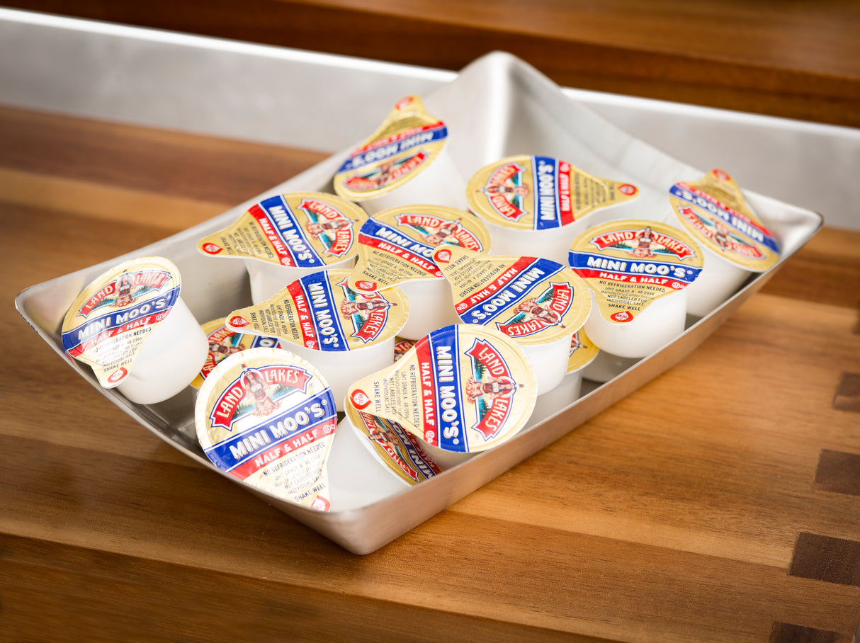 A stainless steel fry tray holds several individual Land O’Lakes Mini Moo’s half & half creamers with blue, red, and yellow labels. The tray sits on a wooden counter with a metal background, and the creamers are scattered and overlapping.