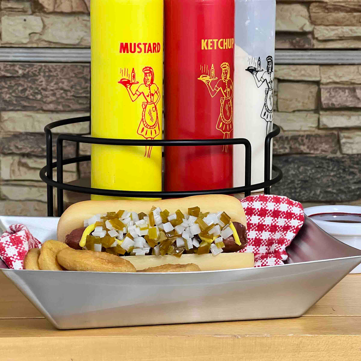 A hot dog topped with chopped onions and relish sits in a stainless steel fry tray beside onion rings. Behind it, large condiment dispensers from the Better Burger Collection add a retro flair against the brick wall background.