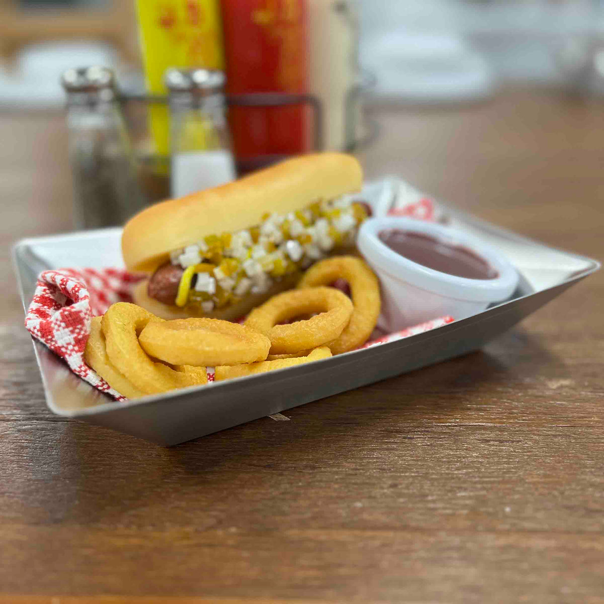 A hot dog topped with diced onions and relish in a bun sits on a fry tray beside golden curly fries and a cup of ketchup. Salt and pepper shakers, plus ketchup and mustard bottles, are blurred in the background of this Better Burger Collection scene.