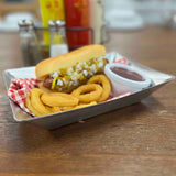 A hot dog topped with diced onions and relish in a bun sits on a fry tray beside golden curly fries and a cup of ketchup. Salt and pepper shakers, plus ketchup and mustard bottles, are blurred in the background of this Better Burger Collection scene.