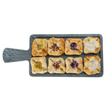 A wooden serving paddle holds eight square pastries, each with a golden, flaky crust and colorful fruit or cream fillings at the center. Lightly drizzled with white glaze, this display pairs beautifully with the Smokehouse Melamine Collection.
