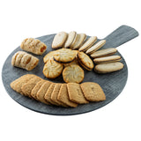 A round, gray serving paddle from the Smokehouse Melamine Collection holds an assortment of cookies: rectangular brown cookies, oval cream-filled cookies, golden rounds, and sugar-dusted pastries. The board is set against a white background.